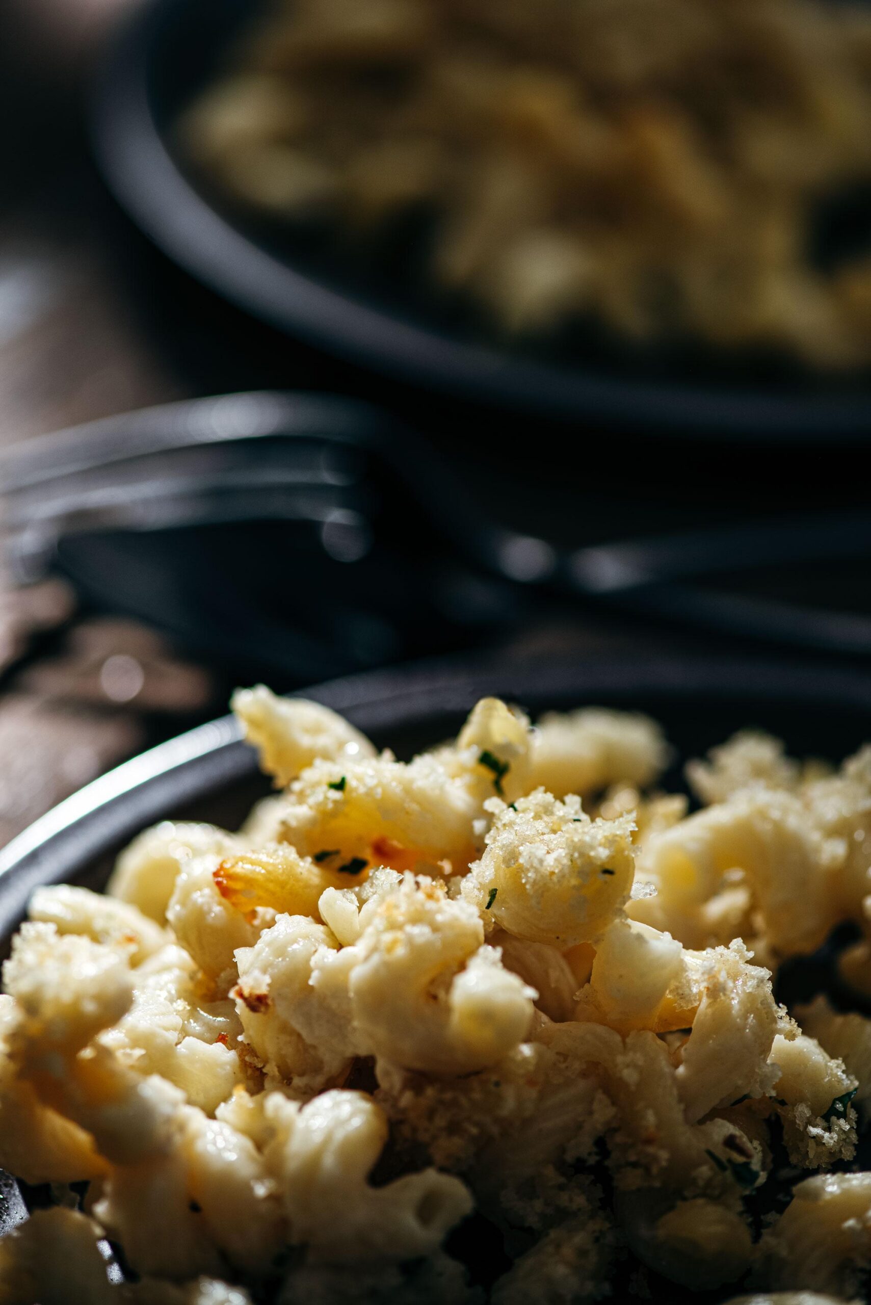 Close-up of baked macaroni and cheese topped with a golden breadcrumb crust, served on a dark plate with another plate and silverware blurred in the background.