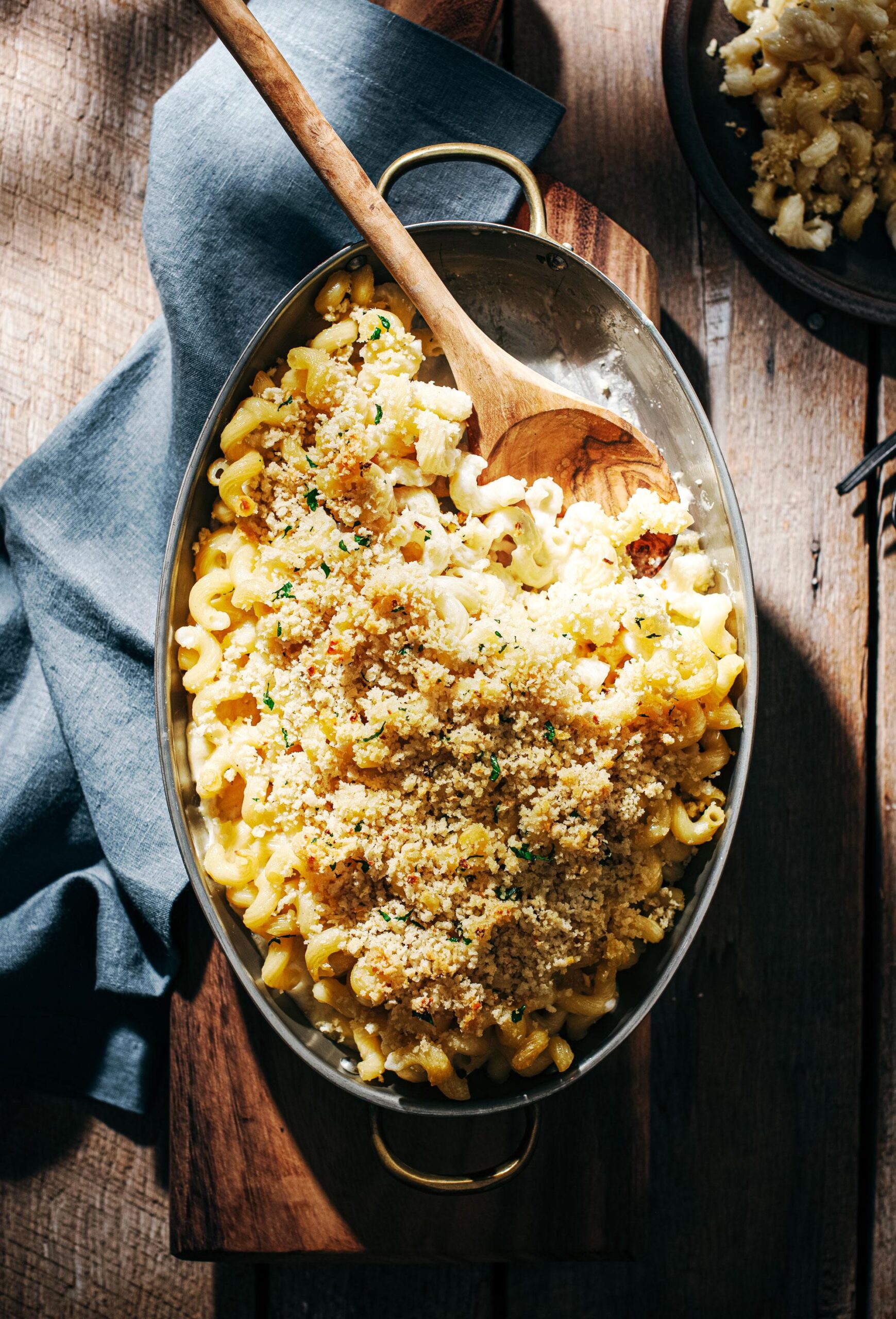 A baking dish of macaroni and cheese topped with golden breadcrumbs, with a wooden spoon resting inside. The dish sits on a wooden surface, partially covered by a blue linen napkin.