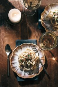 A plate of pasta with mushrooms and herbs is set on a wooden table next to a fork, spoon, a lit candle, and glasses of white wine, creating a cozy and elegant dining atmosphere.