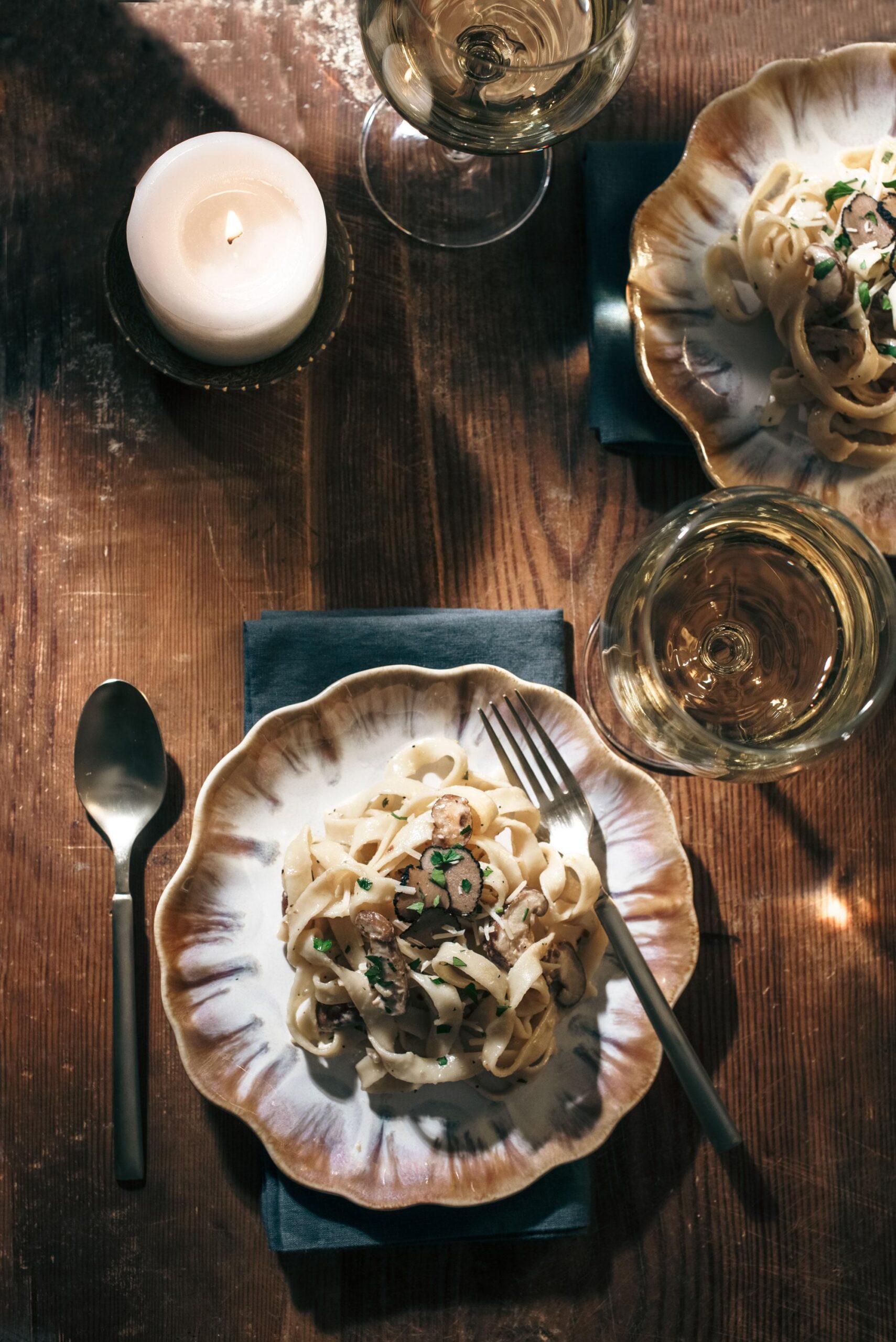 A plate of pasta with mushrooms and herbs is set on a wooden table next to a fork, spoon, a lit candle, and glasses of white wine, creating a cozy and elegant dining atmosphere.
