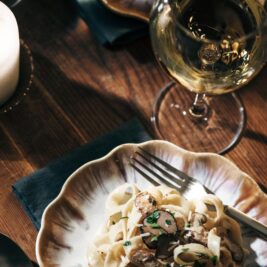 A plate of creamy pasta garnished with herbs sits on a decorative plate beside a fork, with a glass of white wine and another plate of pasta visible in the background on a wooden table.
