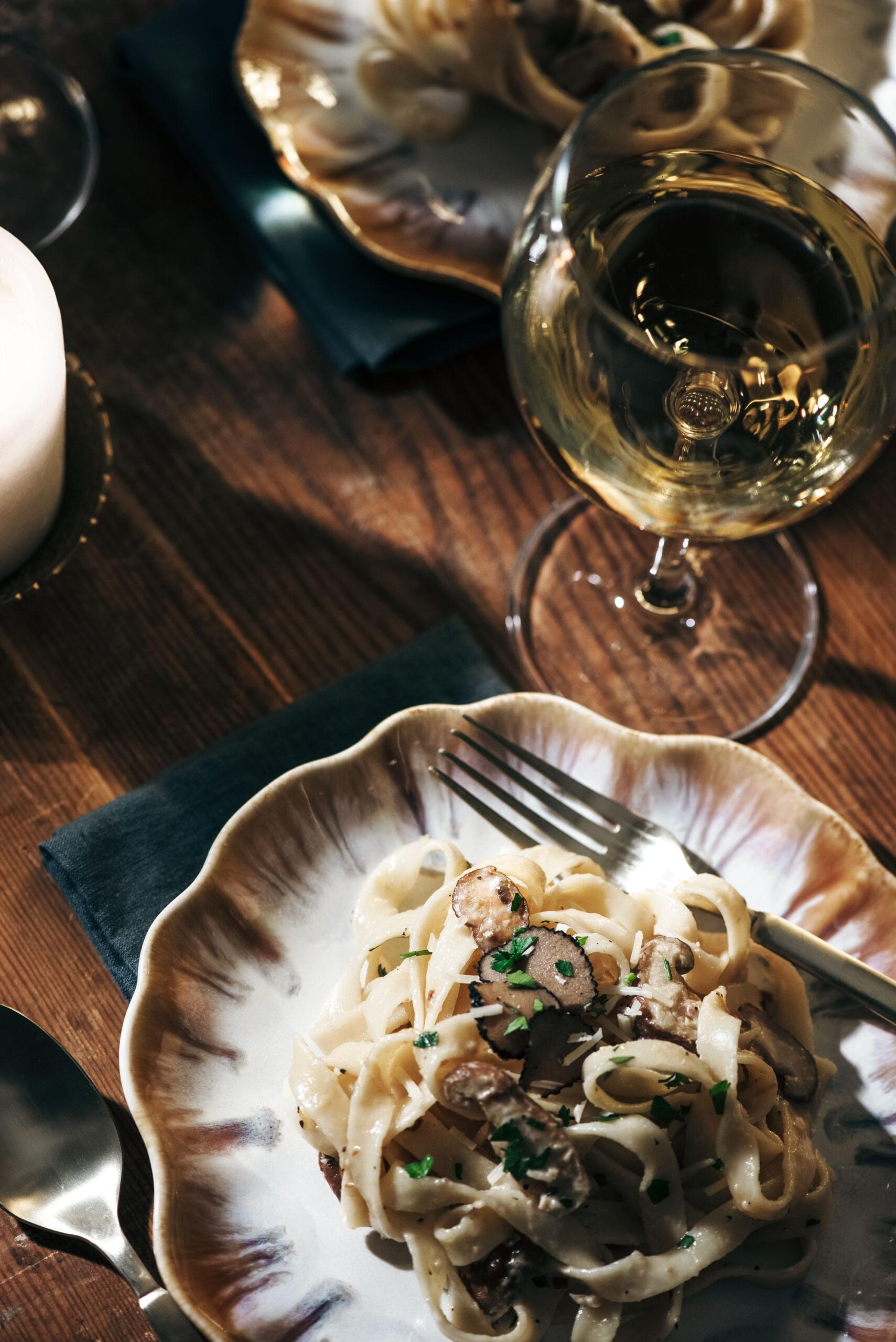 A plate of creamy pasta garnished with herbs sits on a decorative plate beside a fork, with a glass of white wine and another plate of pasta visible in the background on a wooden table.