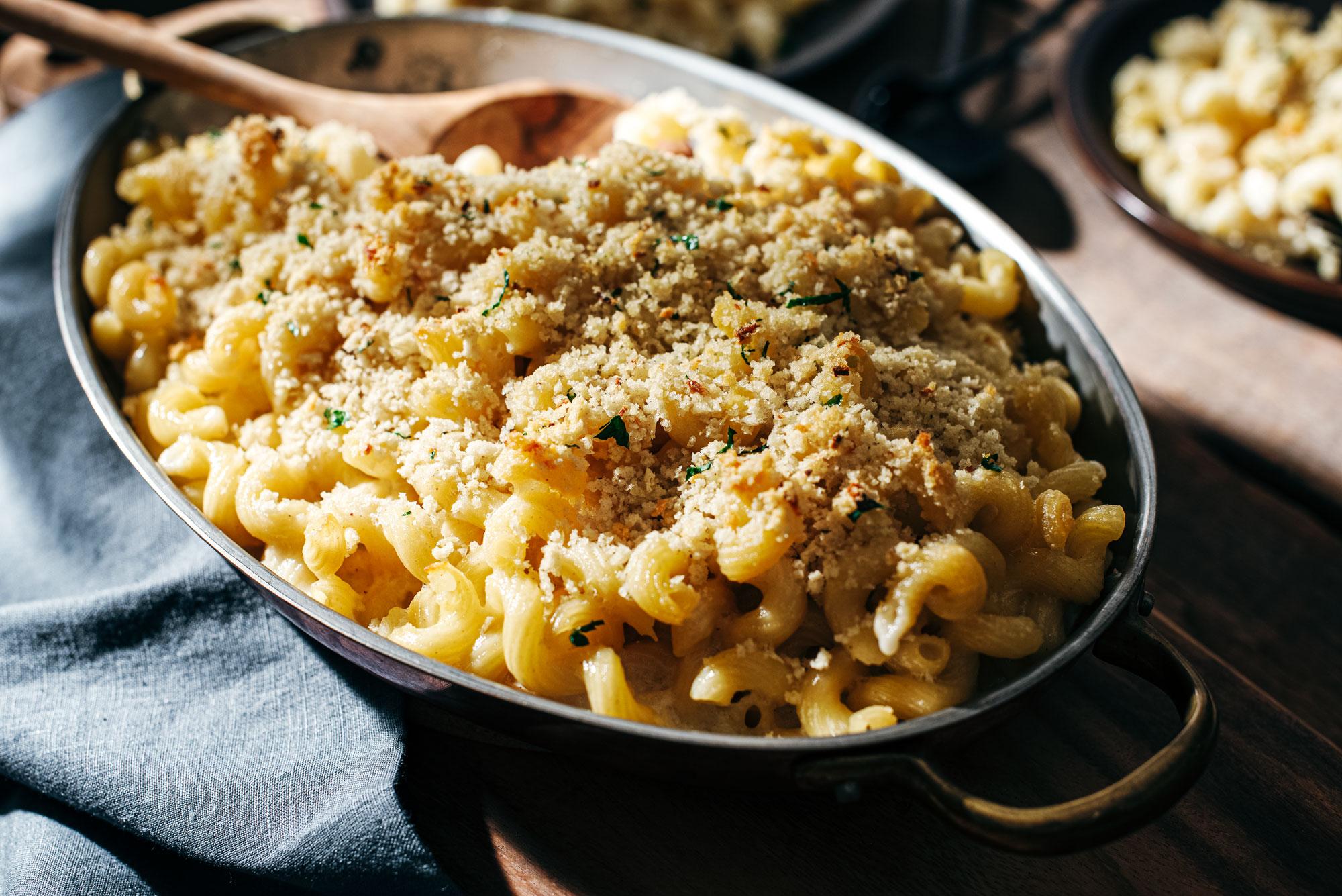 A dish of baked macaroni and cheese topped with golden breadcrumbs sits on a wooden table, with a wooden spoon resting on the side. Soft sunlight highlights the creamy pasta and crisp topping.