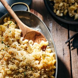 A baking dish of macaroni and cheese topped with golden breadcrumbs, with a wooden spoon resting inside. A plate of the pasta and utensils are visible on a rustic wooden table.