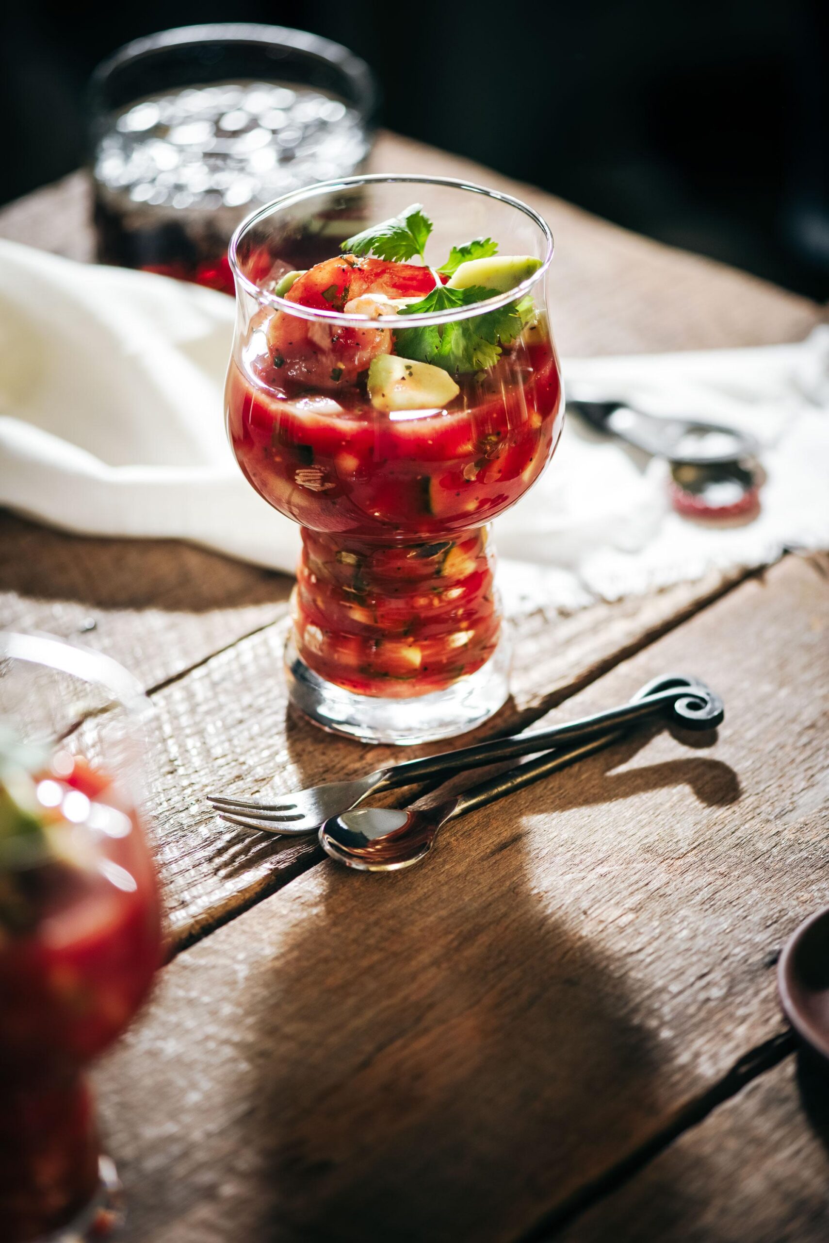 A glass filled with a tomato-based salad garnished with herbs, placed on a rustic wooden table alongside a small fork and spoon. Sunlight highlights the vibrant colors of the dish.
