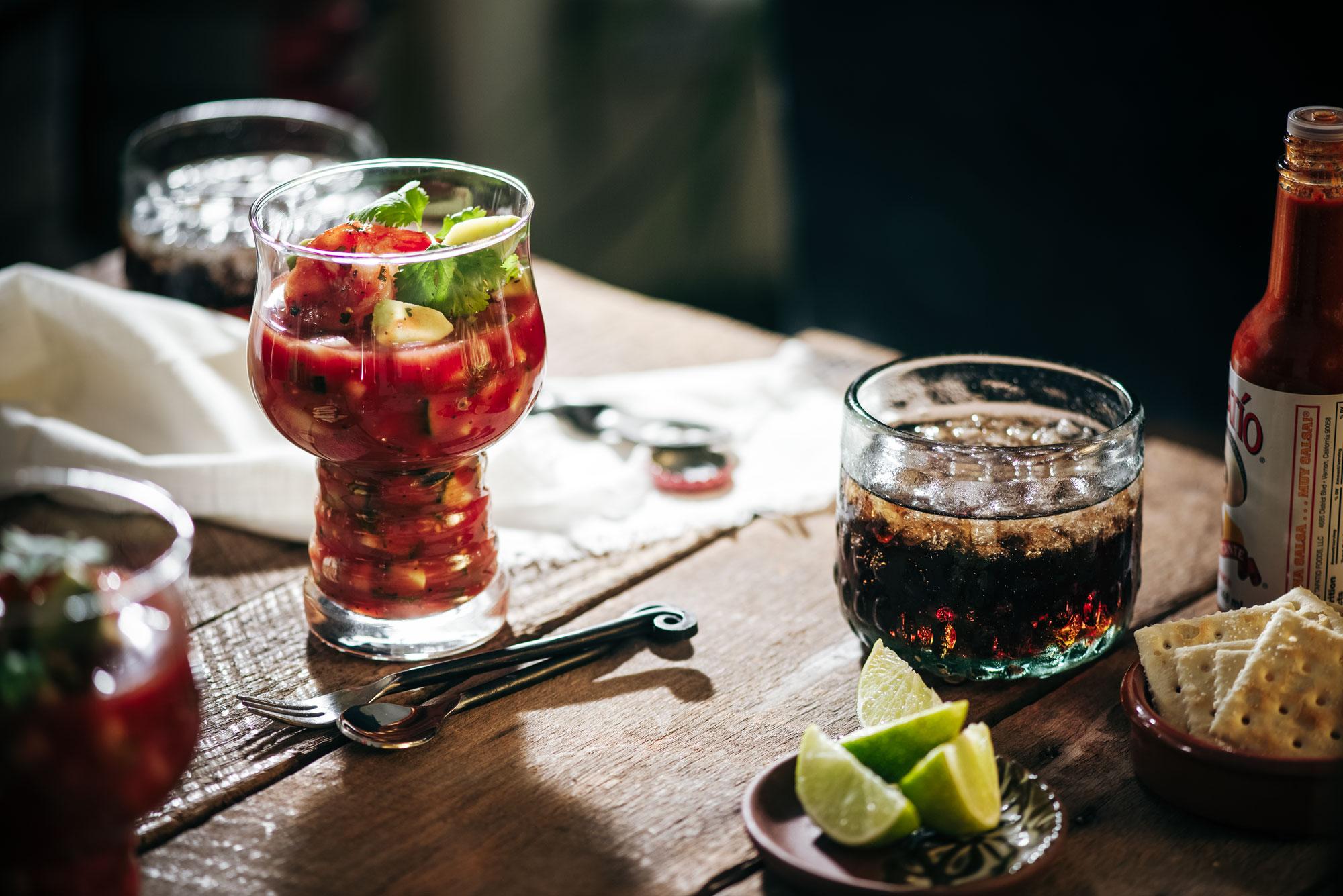 A goblet glass filled with shrimp cocktail garnished with cilantro sits on a wooden table beside a glass of soda, a plate of lime wedges and crackers, and a bottle of hot sauce. Sunlight highlights the scene.