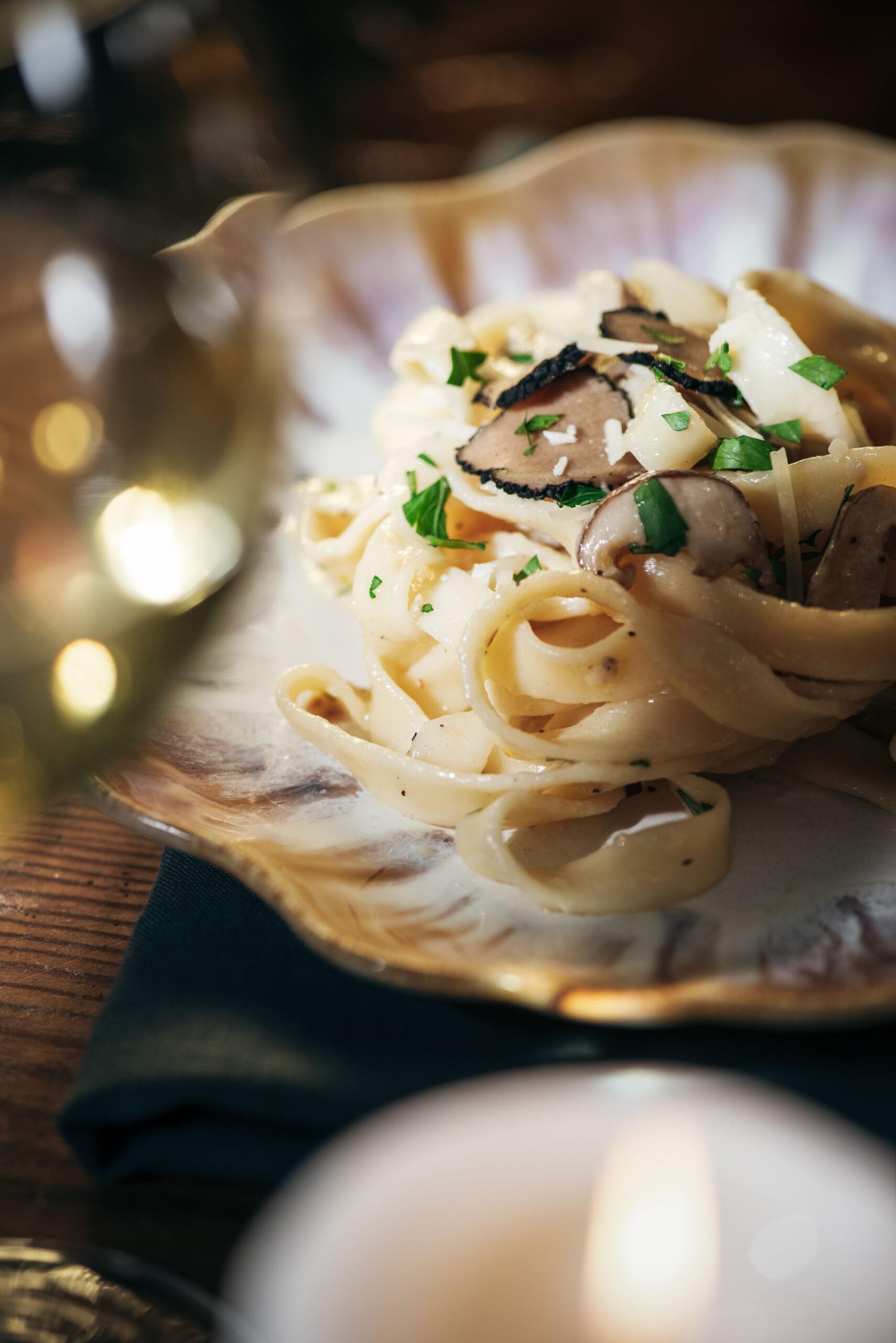 A close-up of a plate of creamy fettuccine pasta garnished with slices of black truffle and chopped parsley, set on a rustic table with a blurred glass and candle in the foreground.