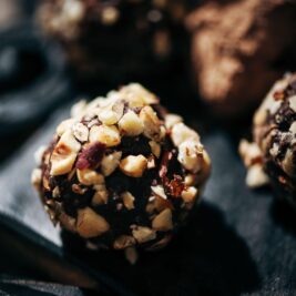 A close-up of a chocolate truffle coated with chopped nuts, resting on a dark surface. The background is softly blurred, showing another truffle lightly dusted with cocoa powder. The texture and detail of the nuts are prominent.