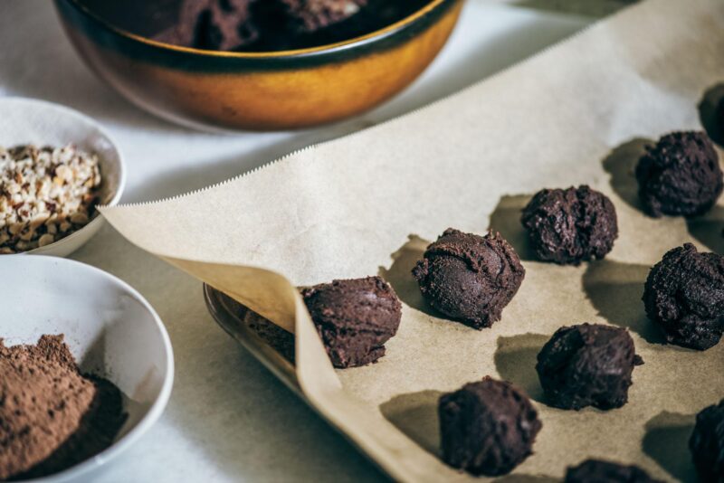Balls of chocolate cookie dough arranged on a baking tray lined with parchment paper. Nearby are bowls containing chopped nuts and cocoa powder. A larger bowl of cookie dough is partially visible in the background.