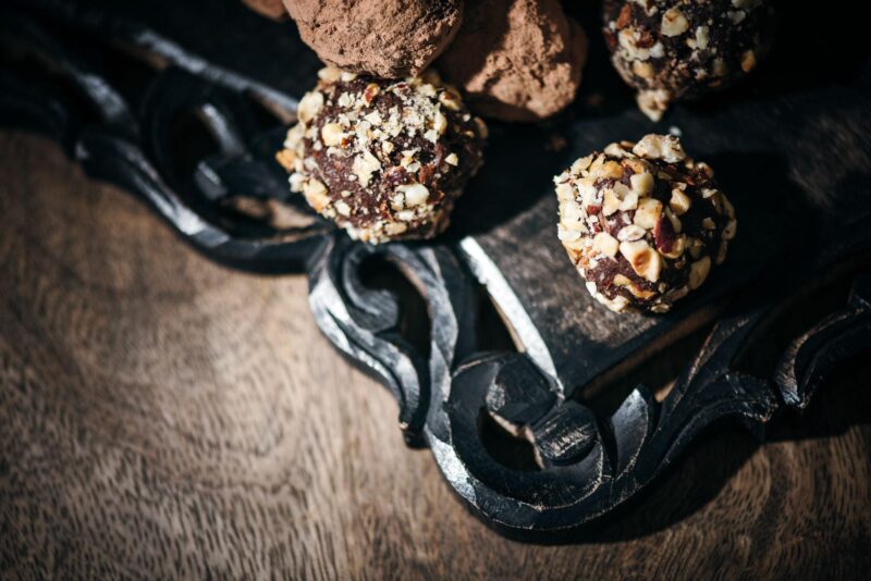Close-up of chocolate truffles covered in chopped nuts and cocoa powder on a dark, ornate wooden tray. The rich texture and warm tones create a luxurious, inviting atmosphere.
