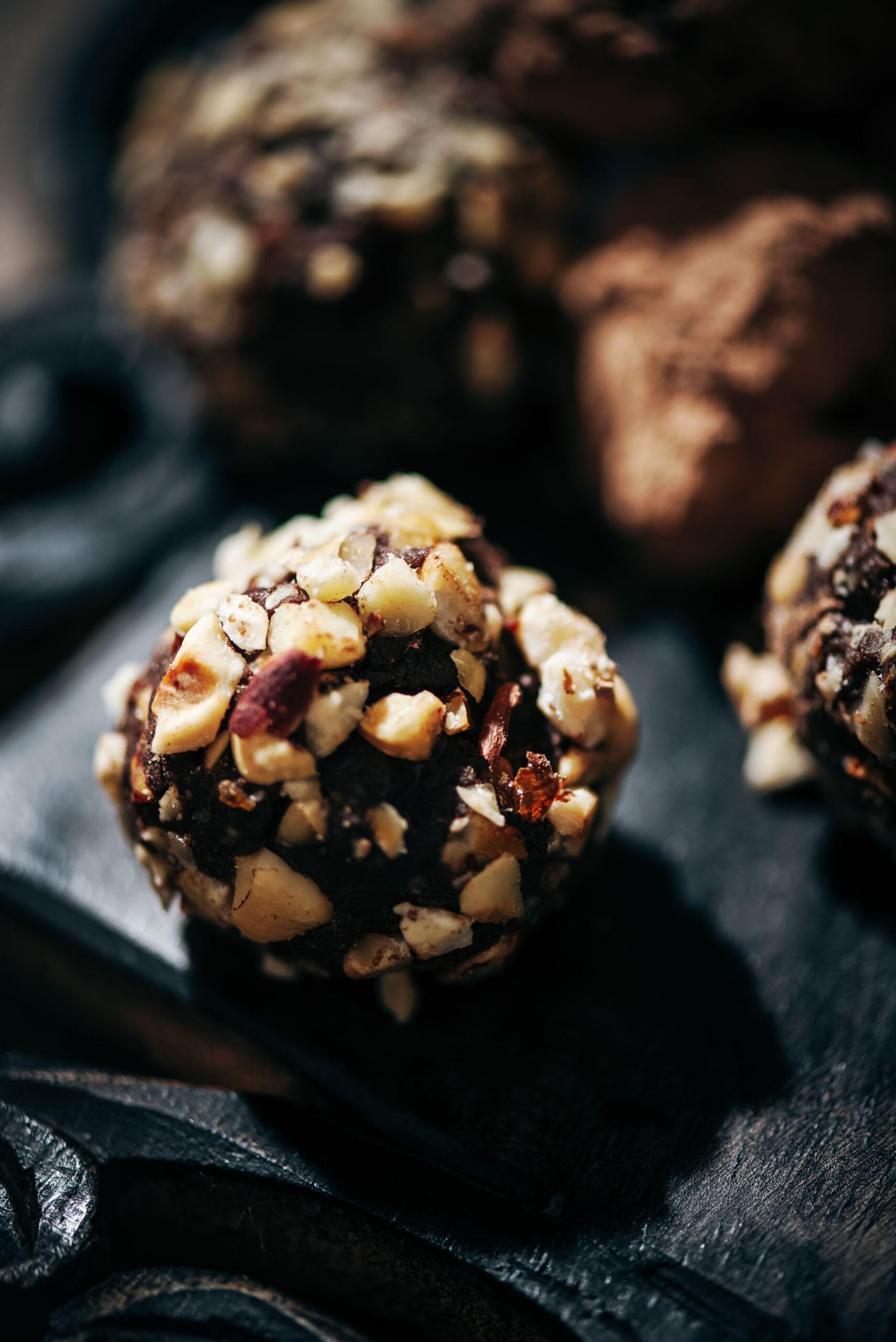 A close-up of a chocolate truffle coated with chopped nuts, resting on a dark surface. The background is softly blurred, showing another truffle lightly dusted with cocoa powder. The texture and detail of the nuts are prominent.
