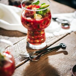 A glass filled with a vibrant tomato and herb salad on a rustic wooden table, accompanied by a spoon and fork. Another glass and a white cloth are in the background. Sunlight casts soft shadows across the scene.