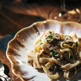 A plate of creamy pasta topped with mushrooms and garnished with herbs. The dish is served on a decorative white and gold-rimmed plate. A fork and knife are nearby, with a glass of white wine blurred in the background.