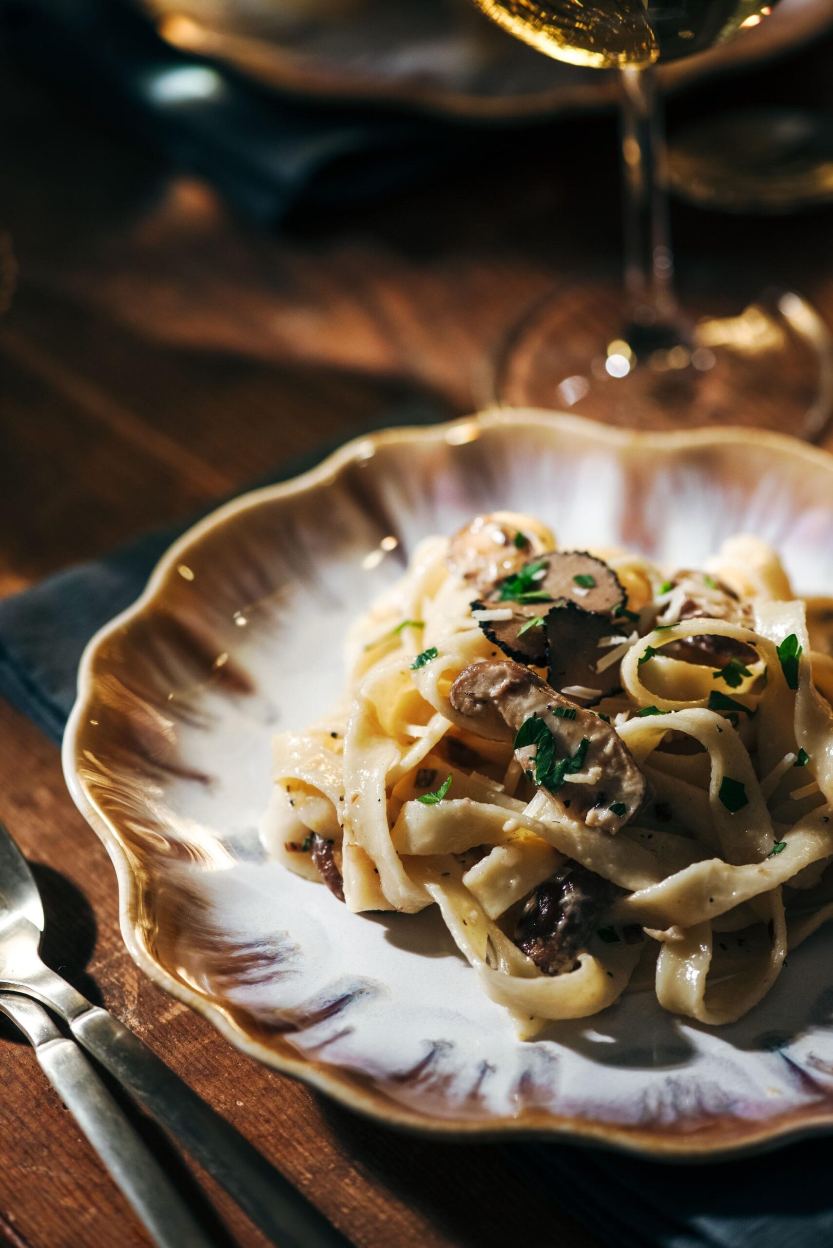 A plate of creamy pasta topped with mushrooms and garnished with herbs. The dish is served on a decorative white and gold-rimmed plate. A fork and knife are nearby, with a glass of white wine blurred in the background.