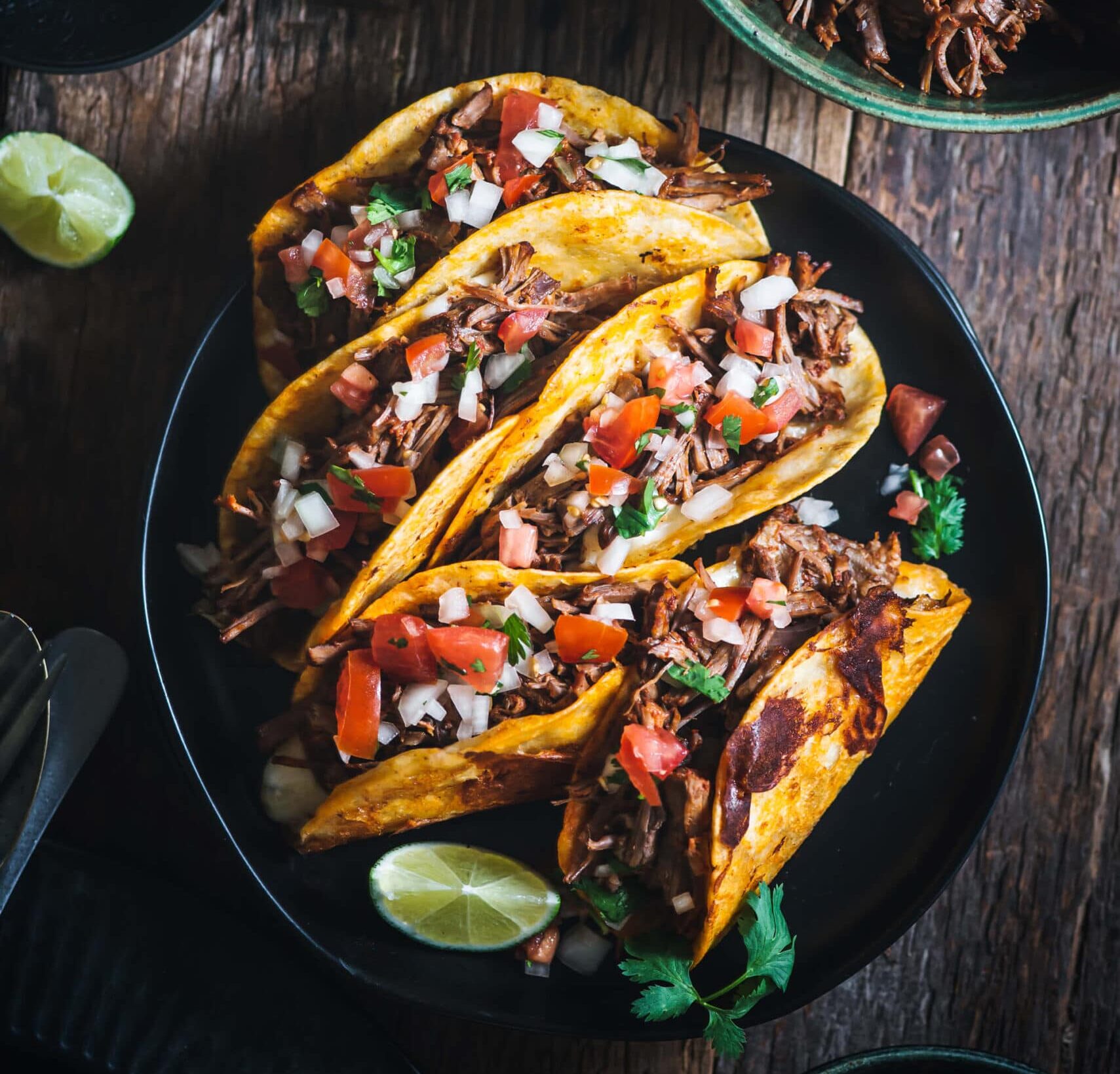 A plate of three tacos filled with shredded meat, diced tomatoes, and onions, garnished with cilantro. There are lime wedges on the plate and a bowl of shredded meat nearby, all set on a rustic wooden table.