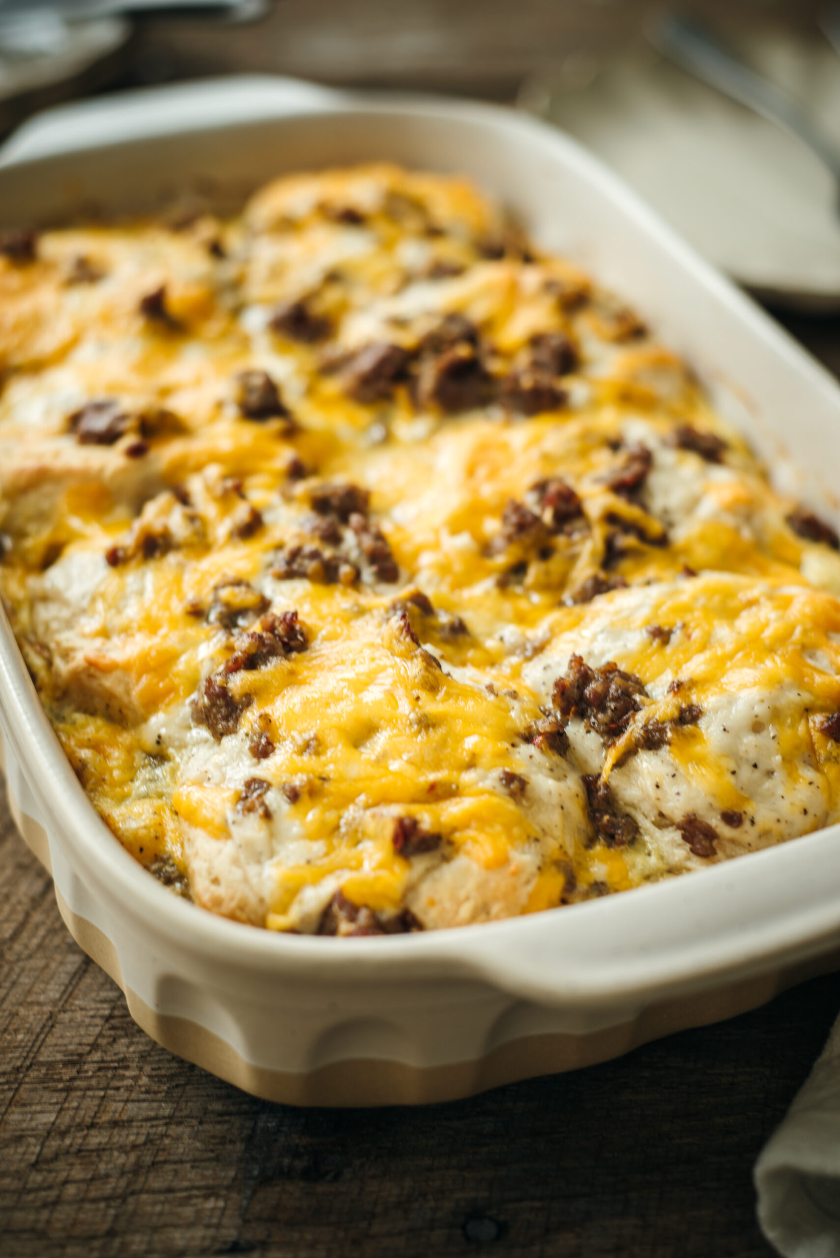 Close-up of a freshly baked sausage biscuit casserole in a white baking dish, topped with melted cheddar cheese and browned sausage pieces on a rustic wooden table