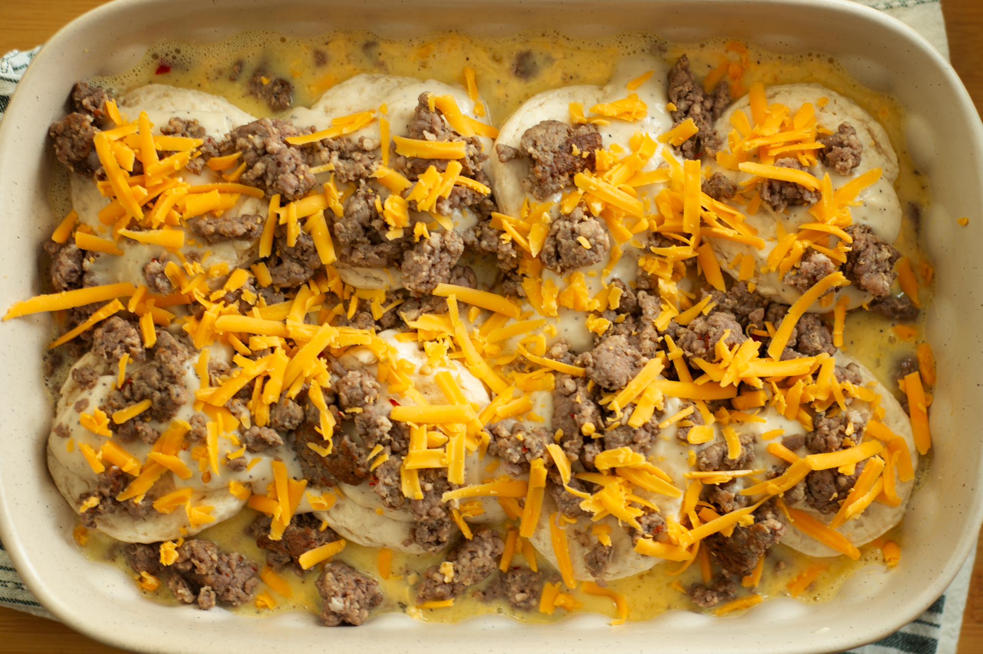 Overhead view of an unbaked sausage biscuit casserole in a white baking dish, layered with biscuits, cooked sausage, shredded cheddar cheese, and creamy gravy ready to go into the oven