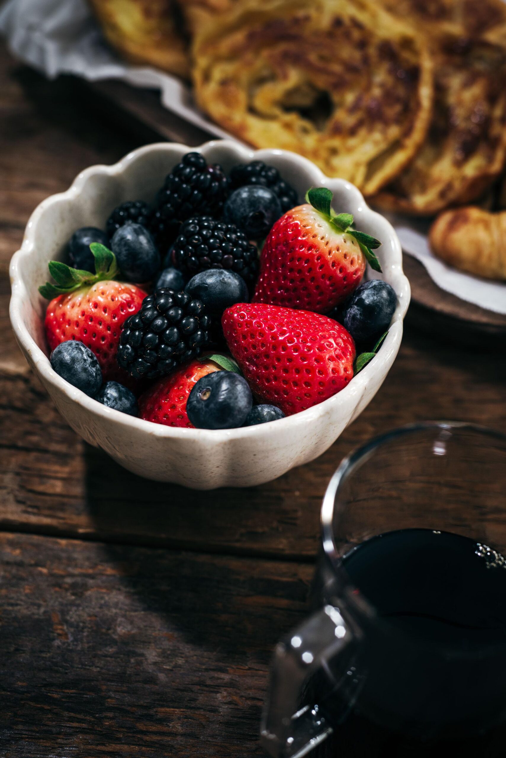 A white bowl filled with strawberries, blueberries, and blackberries sits on a wooden table next to a glass of dark liquid and a plate of pastries in the background.