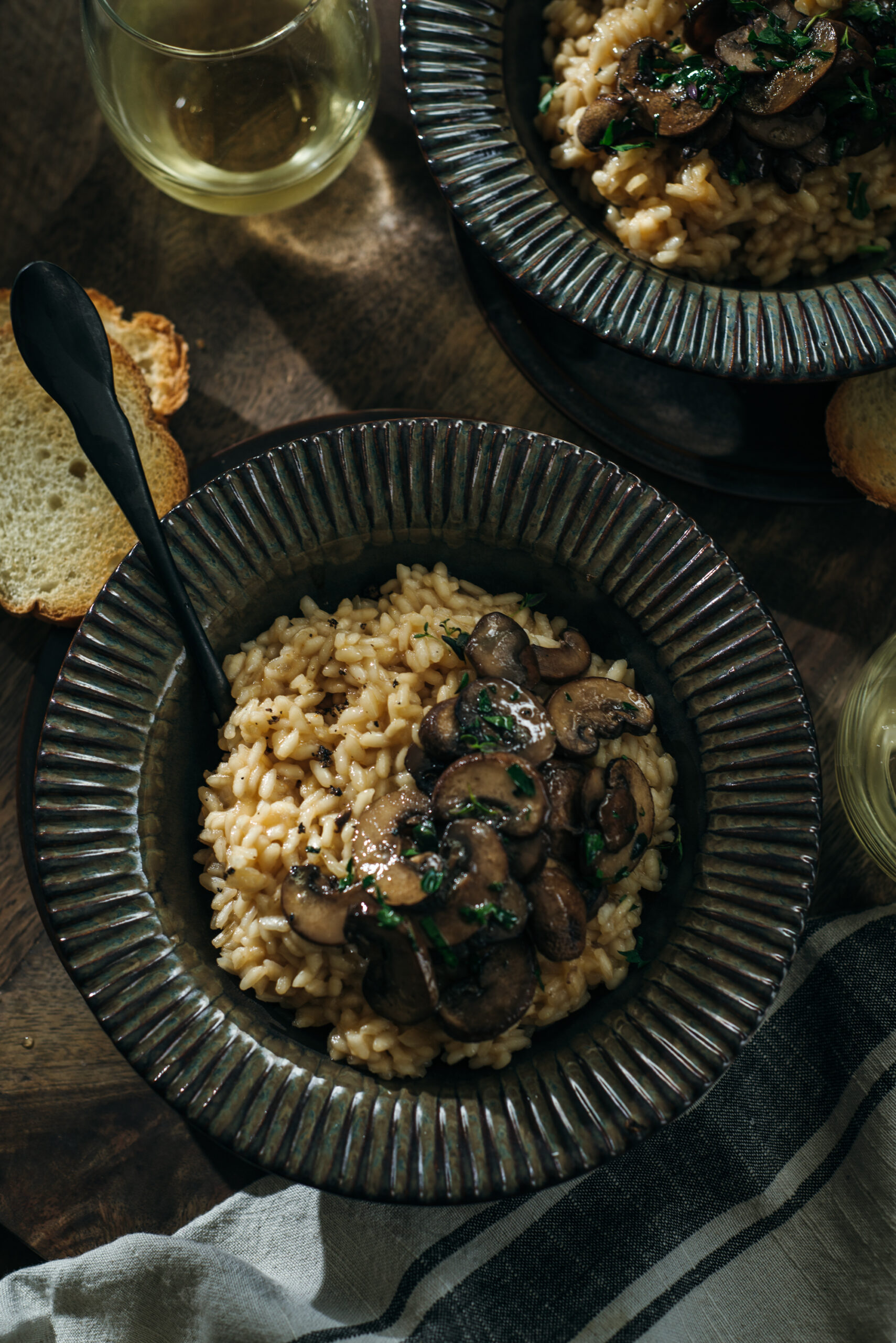 op view of a bowl of mushroom risotto topped with sautéed mushrooms and herbs, served with toasted bread and a glass of white wine on a wooden table