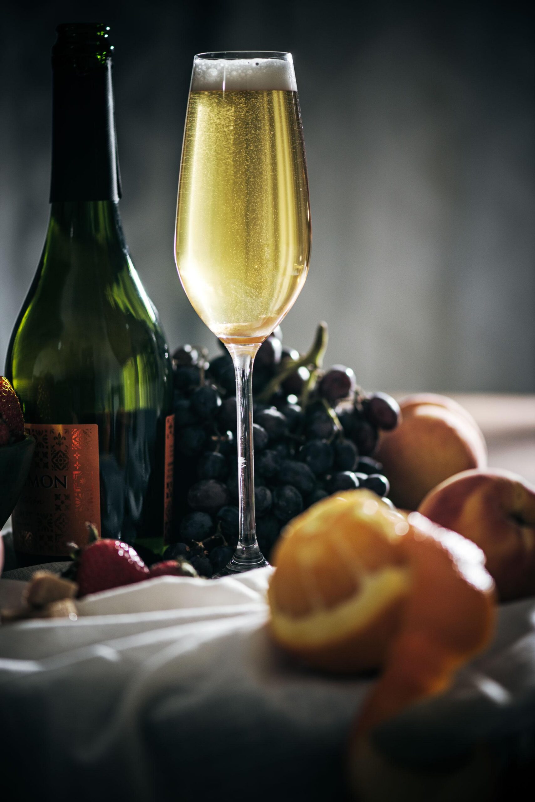 A glass of sparkling wine sits next to a green wine bottle, with a bunch of grapes, strawberries, peaches, and a peeled orange arranged on a table in soft, moody lighting.