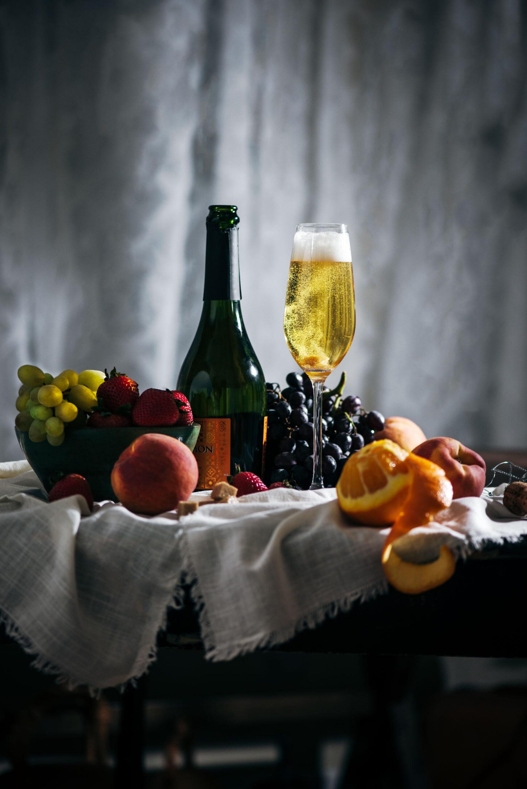 A bottle of sparkling wine, a filled flute, assorted fruits including grapes, strawberries, peaches, and an orange, arranged on a fabric-covered table with soft, moody lighting.