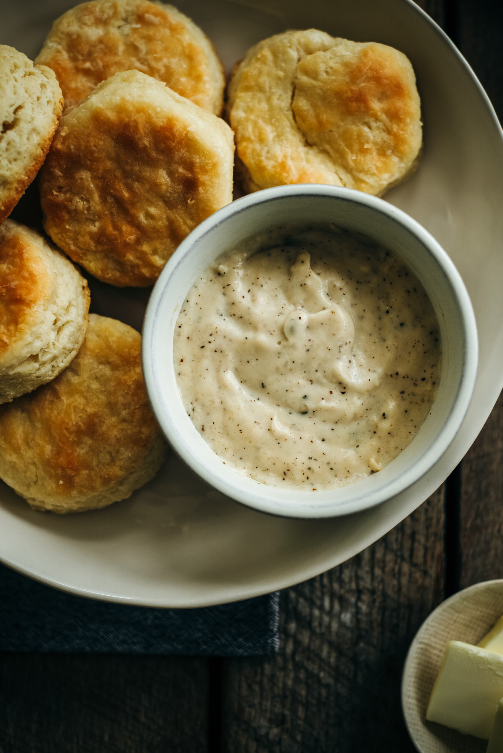 Close-up overhead view of flaky biscuits on a plate beside a bowl of creamy peppered gravy.