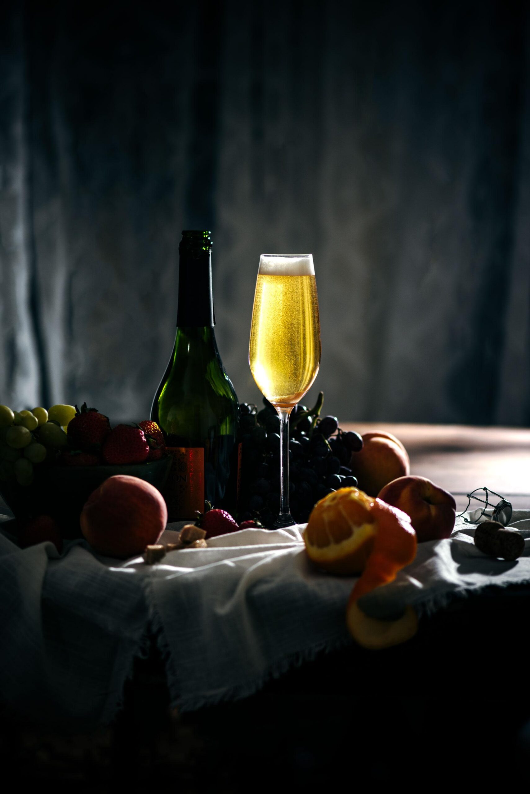 A glass of champagne stands next to a green bottle on a table with peaches, grapes, strawberries, walnuts, and a peeled orange, all arranged on a white cloth in dim lighting.