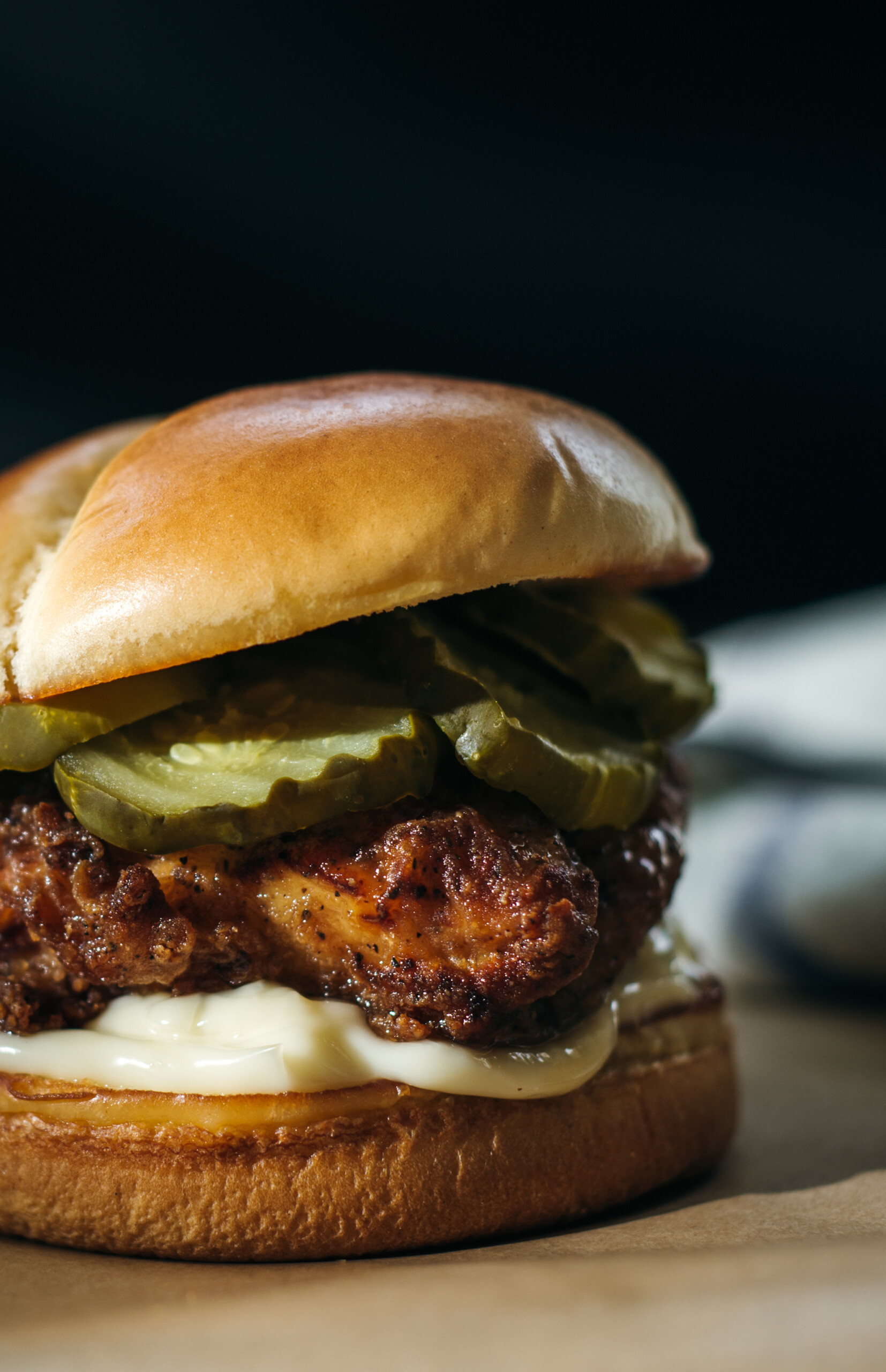 A close-up side view of a crispy fried chicken sandwich topped with pickles and creamy sauce on a toasted bun, set against a dark background.