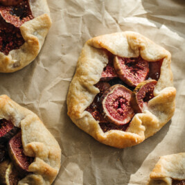 Rustic mini fig galettes with golden-brown crusts and sliced figs baked in the center, drizzled with honey and arranged on parchment paper.