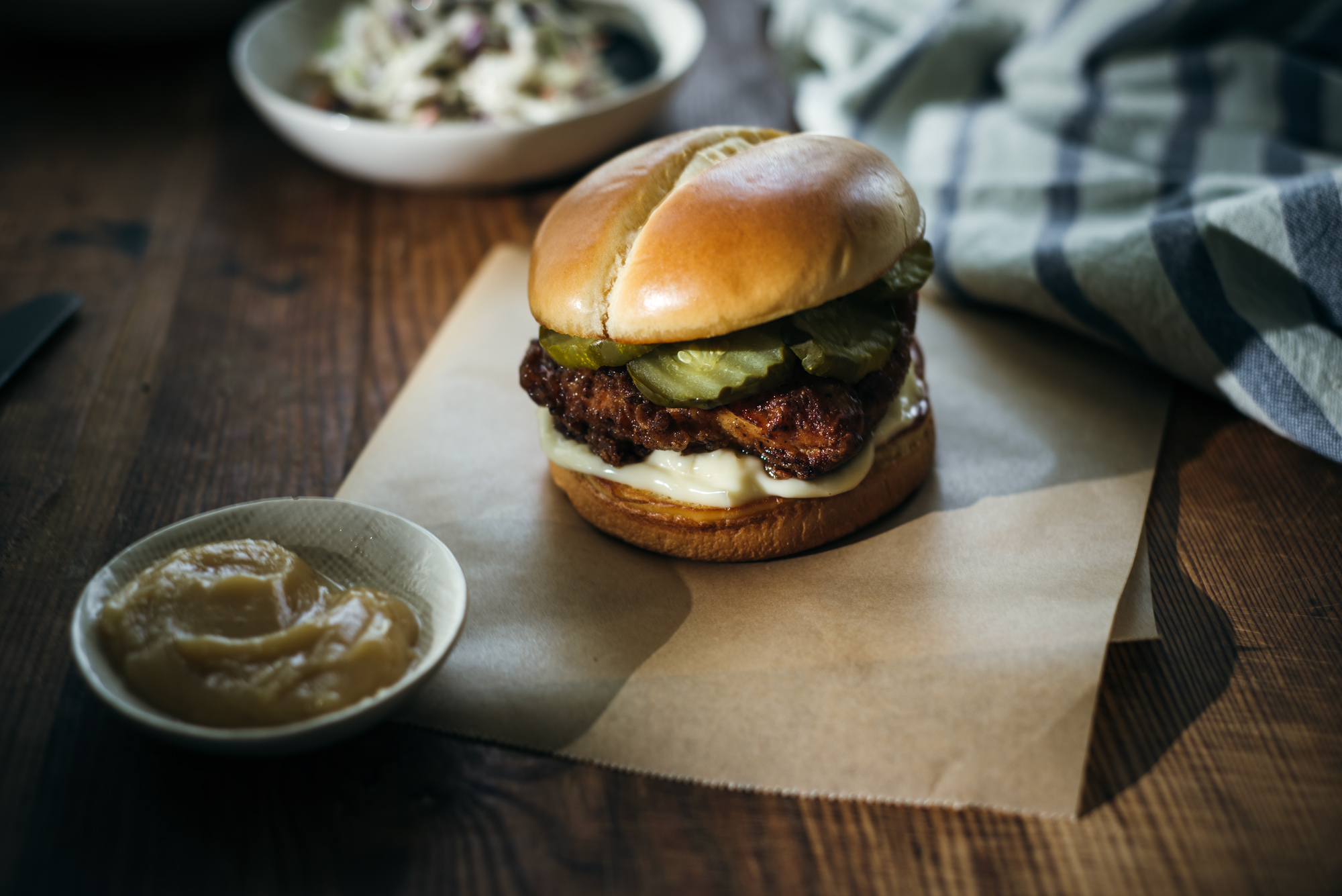 A honey butter chicken sandwich with pickles and creamy sauce on a golden bun sits on parchment paper, with a bowl of coleslaw and honey butter blurred in the background on a rustic table.