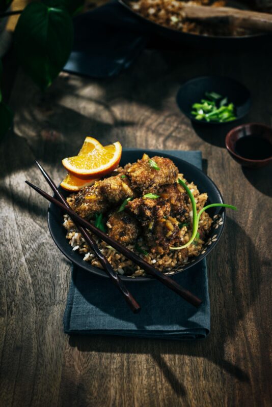 A dark bowl filled with crispy fried chicken atop rice, garnished with green onions, orange slices, and chopsticks, sits on a dark napkin on a wooden table.
