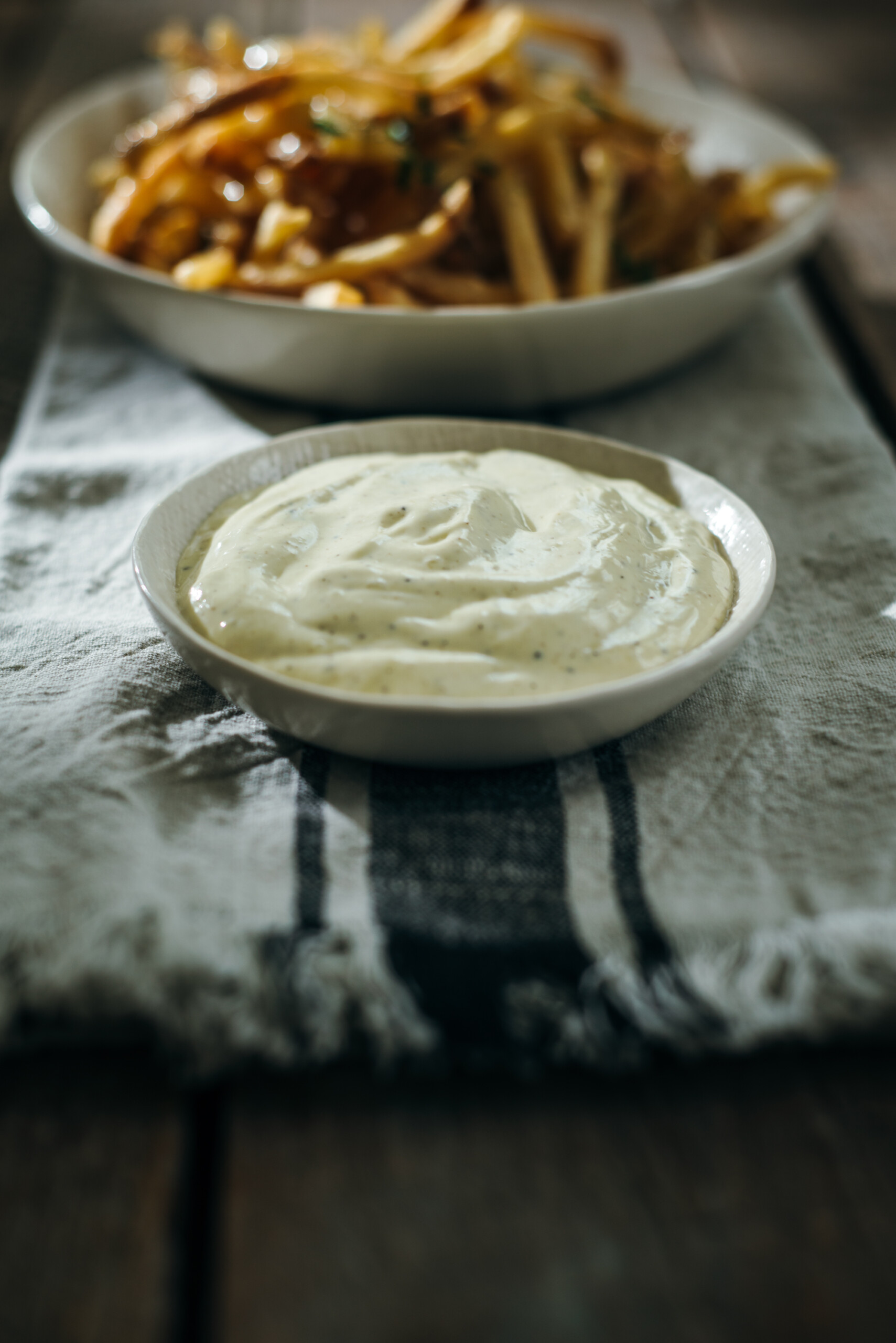 A close-up of a small white bowl filled with creamy truffle aioli placed on a striped cloth, with a bowl of golden fries blurred in the background on a rustic wooden table.