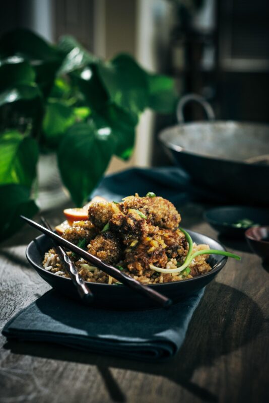A bowl of fried rice topped with crispy fried chicken pieces and vegetables, garnished with green onions. Black chopsticks rest on the bowl, and a leafy plant is blurred in the background.