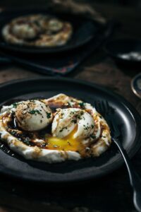 A dark plate holds two poached eggs with runny yolks atop a swirl of creamy yogurt, sprinkled with herbs and spices. A fork rests beside the eggs, and another similar plate is seen blurred in the background.