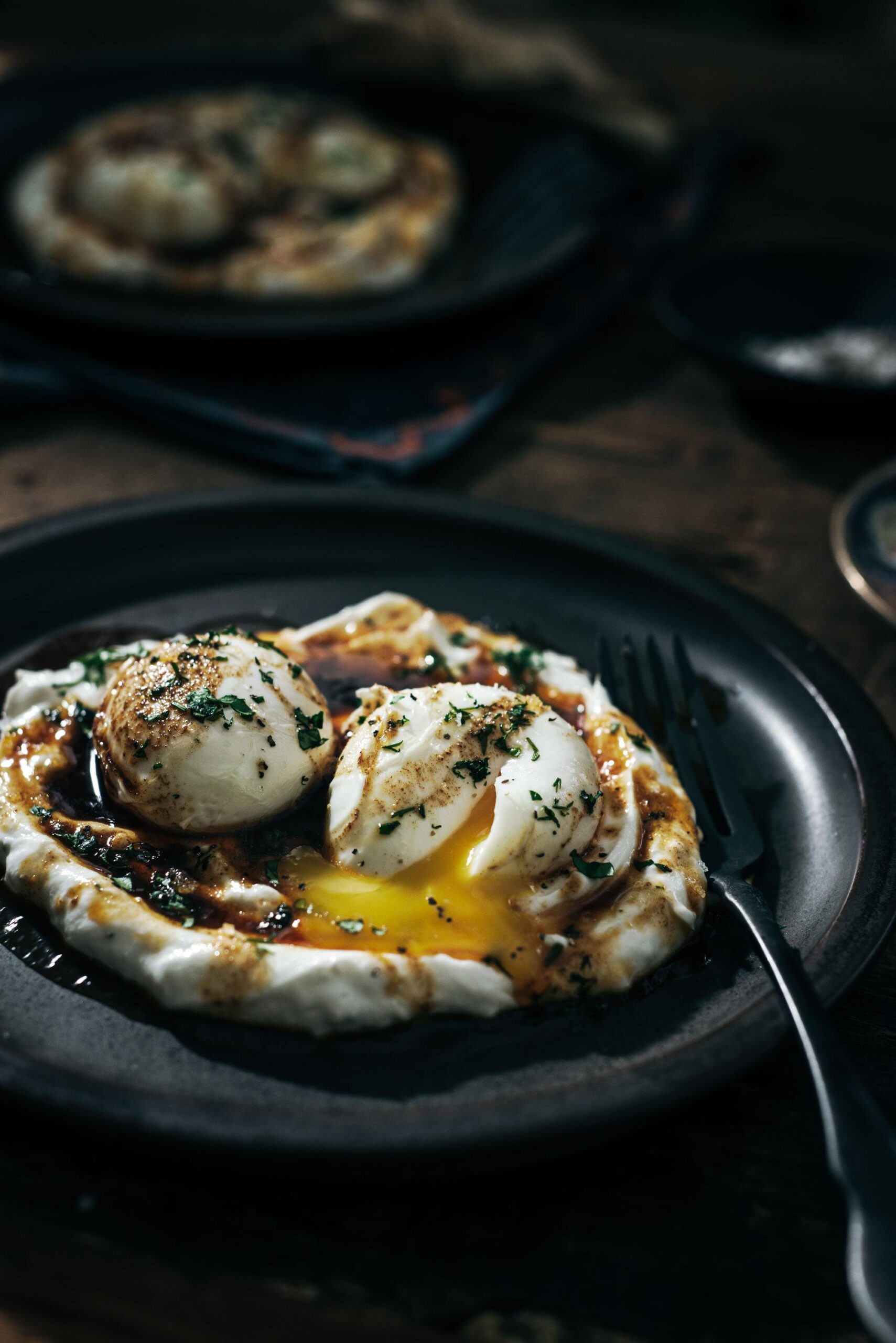 A dark plate holds two poached eggs with runny yolks atop a swirl of creamy yogurt, sprinkled with herbs and spices. A fork rests beside the eggs, and another similar plate is seen blurred in the background.