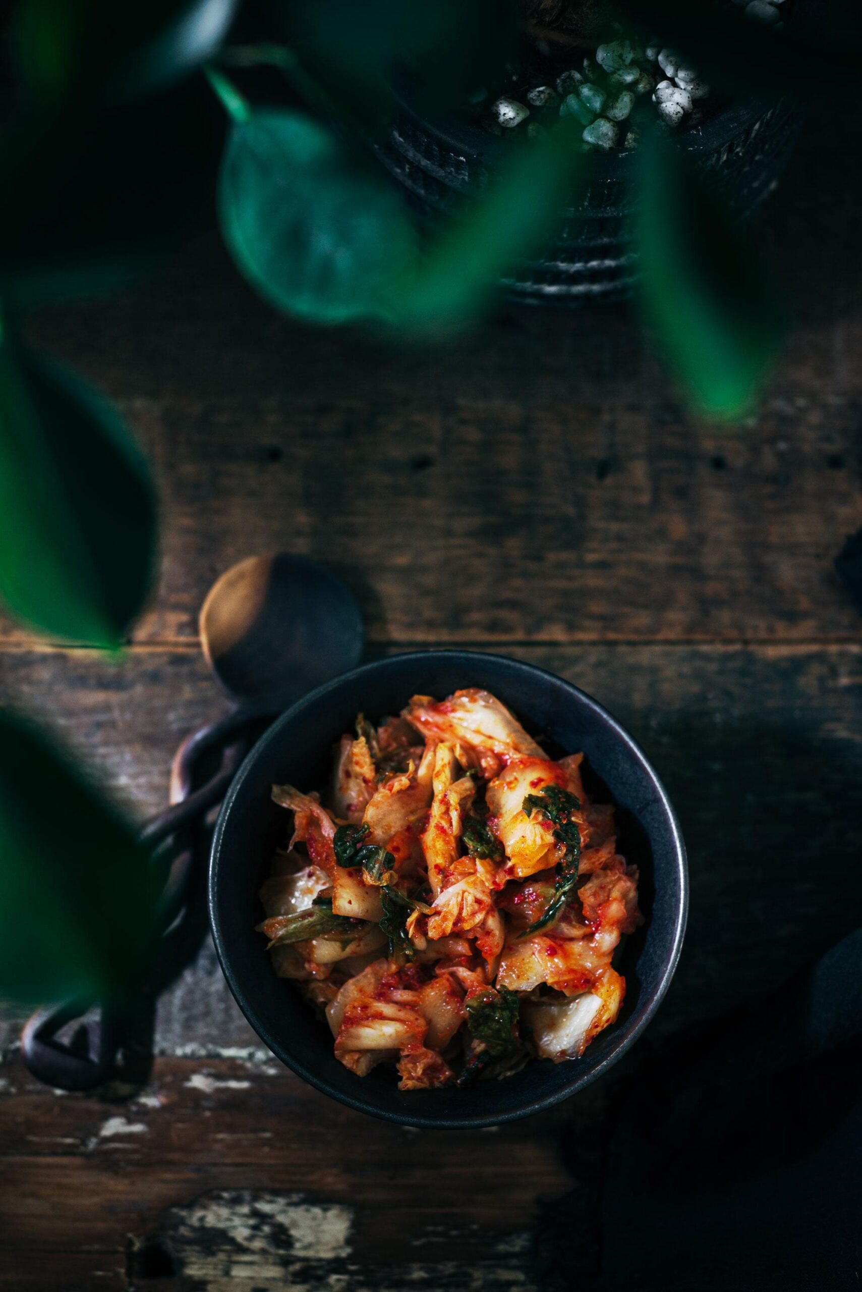 A dark ceramic bowl filled with spicy, red kimchi sits on a wooden table with a wooden spoon beside it. Green leaves partially frame the top of the image, creating a moody atmosphere.
