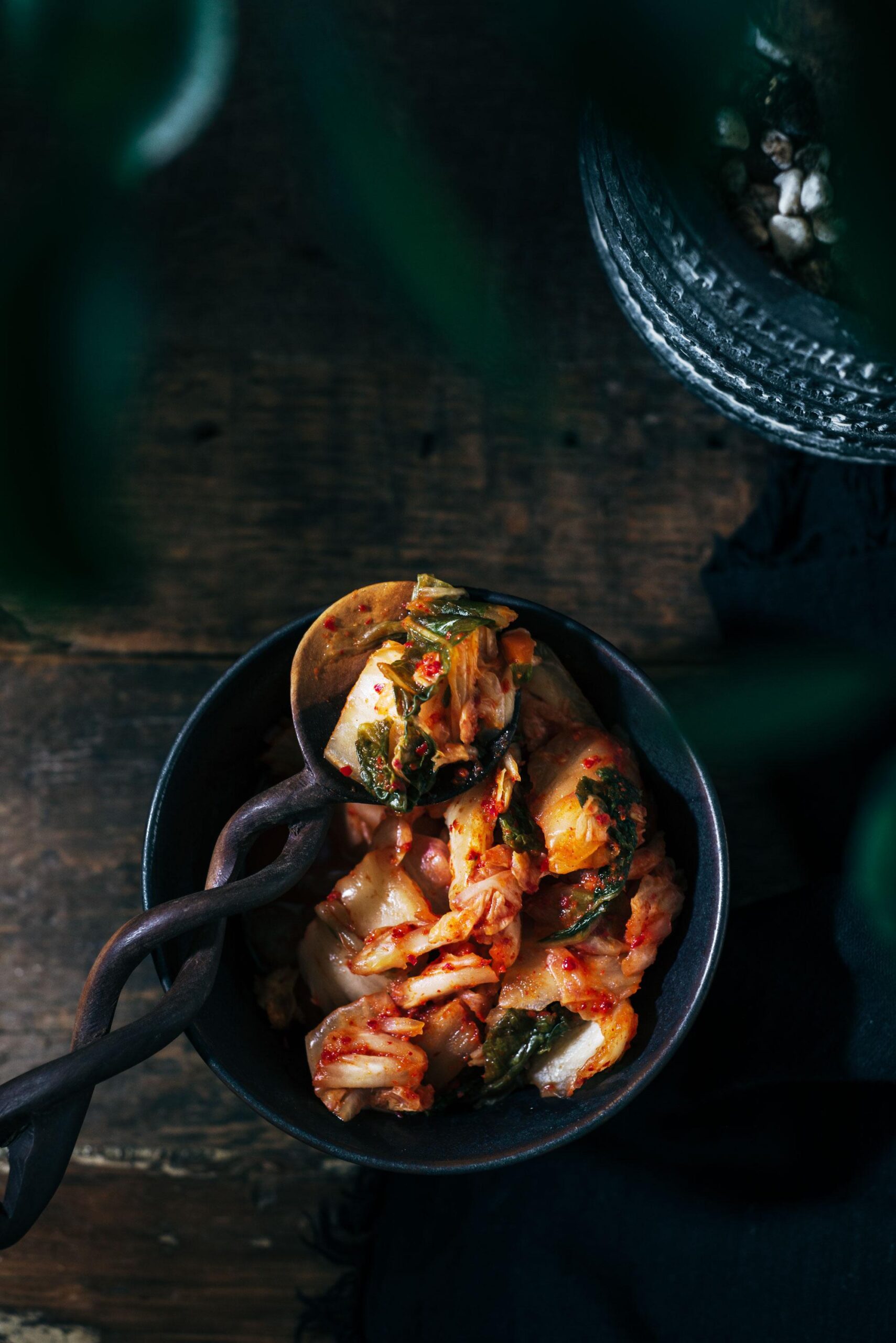 A bowl of kimchi with leafy vegetables is served on a wooden table. A rustic spoon rests in the bowl, and green leaves partially frame the top left of the image, adding depth and a natural touch.