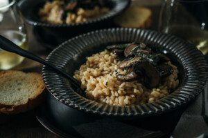 A bowl of creamy mushroom risotto topped with sautéed mushrooms and herbs, served with slices of toasted bread and a glass of white wine on the side