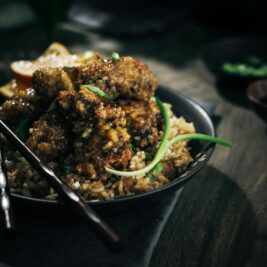 A dark, moody photo of crispy fried chicken pieces coated in sauce served on a bed of fried rice, garnished with green onions. Chopsticks rest on the plate, and a lemon wedge is visible in the background.