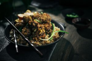 A dark, moody photo of crispy fried chicken pieces coated in sauce served on a bed of fried rice, garnished with green onions. Chopsticks rest on the plate, and a lemon wedge is visible in the background.