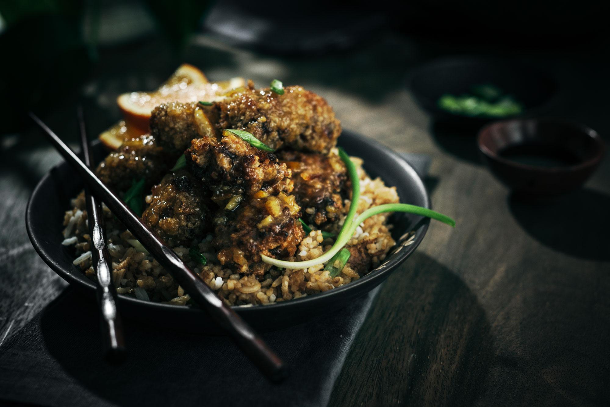 A dark, moody photo of crispy fried chicken pieces coated in sauce served on a bed of fried rice, garnished with green onions. Chopsticks rest on the plate, and a lemon wedge is visible in the background.