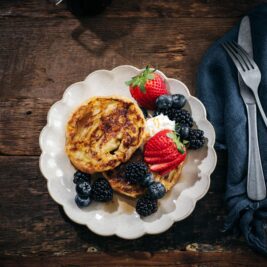 A plate of pancakes topped with whipped cream, fresh strawberries, blackberries, and blueberries sits on a rustic wooden table next to a dark blue napkin and silver cutlery.