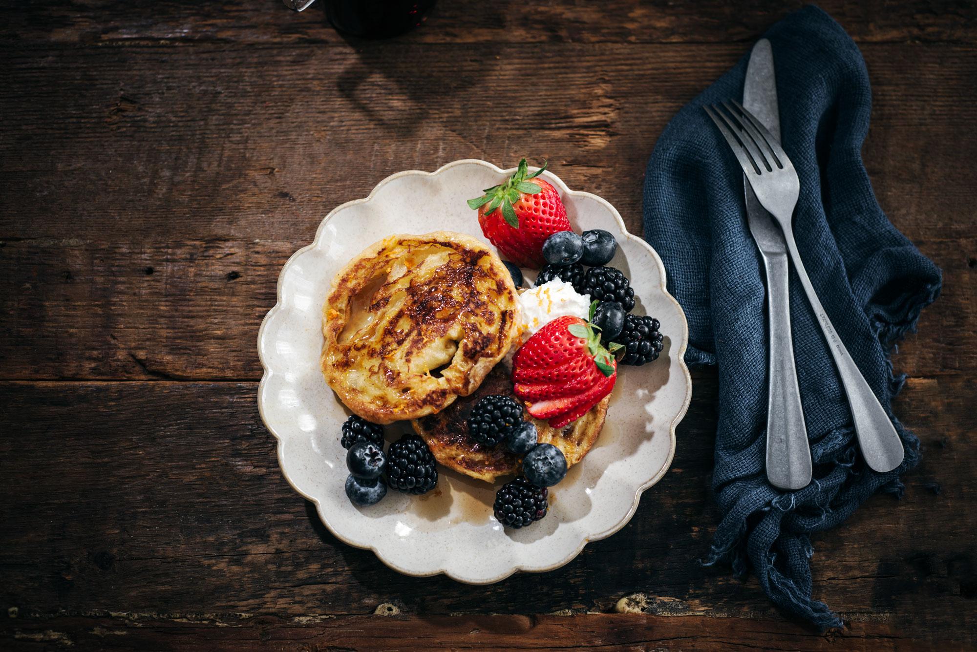 A plate of pancakes topped with whipped cream, fresh strawberries, blackberries, and blueberries sits on a rustic wooden table next to a dark blue napkin and silver cutlery.