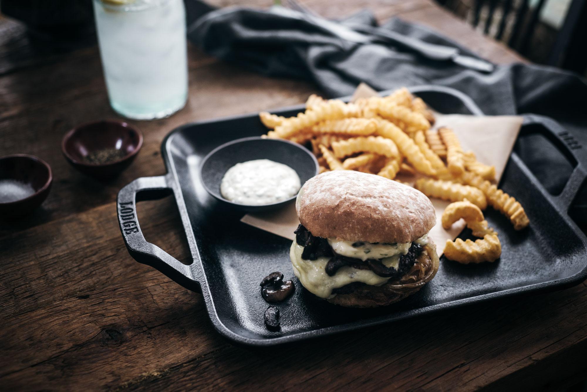 A tray with a cheeseburger topped with sautéed mushrooms, crinkle-cut French fries, and a small dish of dipping sauce on a rustic wooden table. A glass of lemonade is in the background.