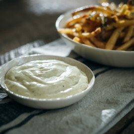 A small white bowl filled with creamy truffle aioli sits on a striped cloth napkin, with a bowl of golden French fries in the background on a rustic wooden table