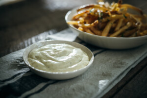 A small white bowl filled with creamy truffle aioli sits on a striped cloth napkin, with a bowl of golden French fries in the background on a rustic wooden table