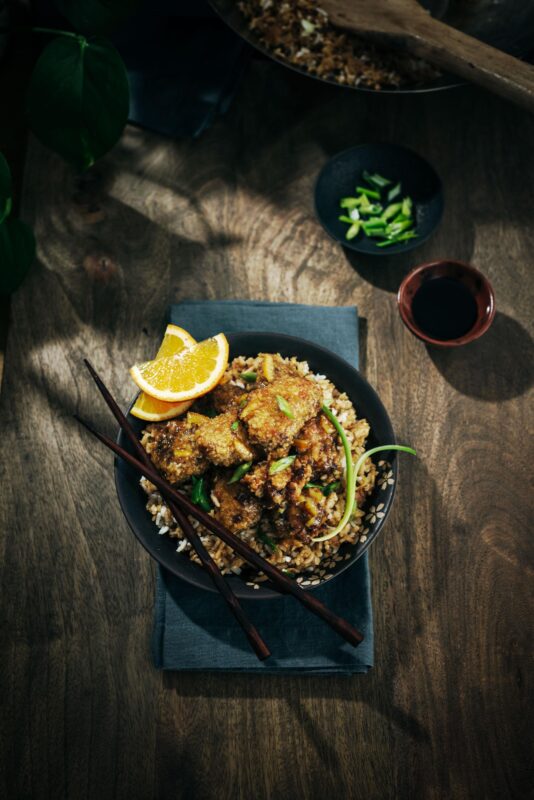 A bowl of fried rice topped with crispy orange chicken, garnished with green onions and an orange slice, with chopsticks resting on the bowl. A small bowl of soy sauce and chopped green onions are nearby on a wooden table.
