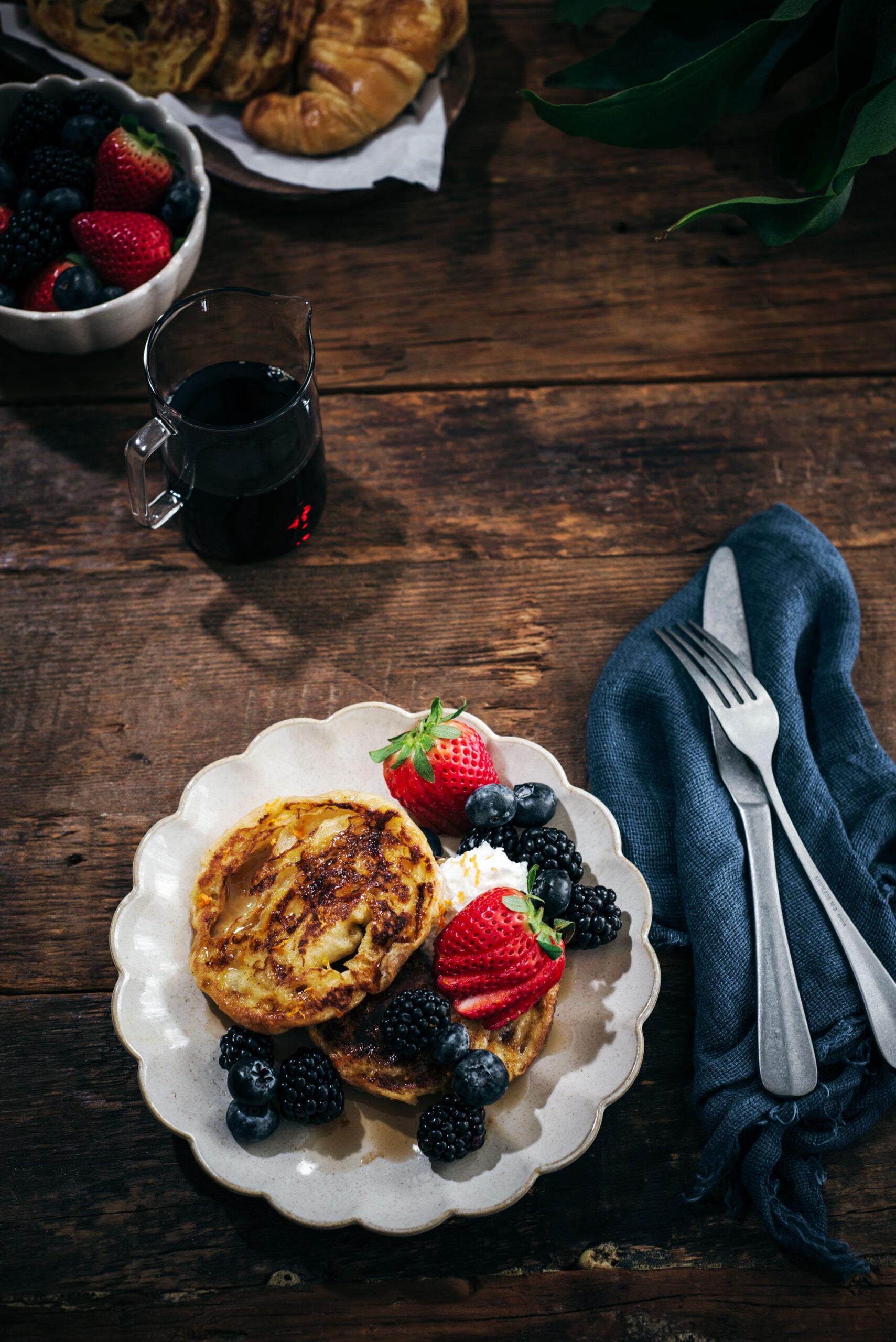 A plate with two pieces of French toast topped with whipped cream, strawberries, blackberries, and blueberries sits on a wooden table next to a fork, knife, napkin, a bowl of berries, and a glass of dark liquid.
