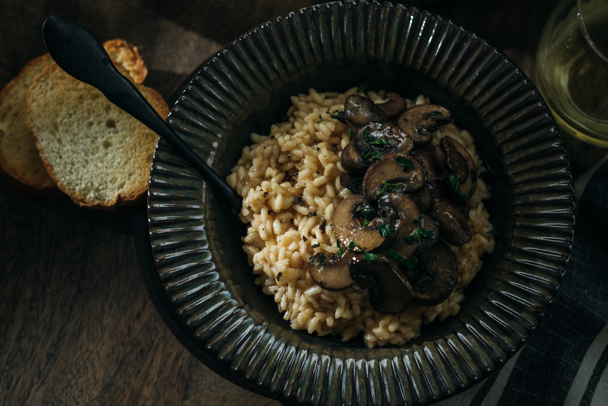 Overhead view of a bowl of mushroom risotto topped with sautéed mushrooms and herbs, served with toasted bread slices and a glass of white wine on a wooden table