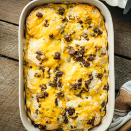 Overhead view of a baked sausage biscuit casserole in a white dish, topped with melted cheddar cheese and browned sausage, resting on a rustic wooden table with a serving spatula nearby